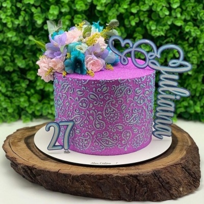 Bolo rosa com flores e decoração prateada, base branca e prato de madeira rústico