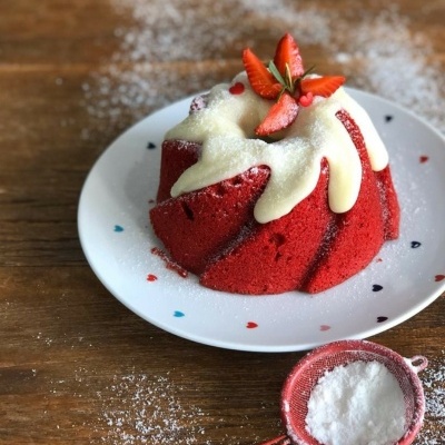 Bolo vermelho tulipa com calda branca e morangos em prato branco com corações, açúcar em coador vermelho sobre mesa de madeira.