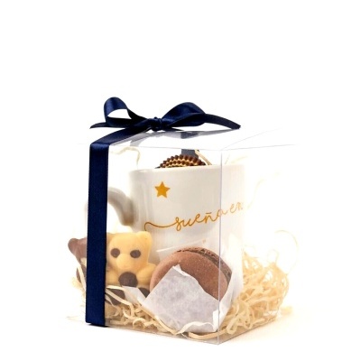 Conjunto de presente com caneca branca, biscoito em forma de urso e bola decorativa numa caixa transparente com fita azul.