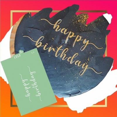 Bolo preto com inscrição dourada 'happy birthday' e cartão verde com a mesma mensagem num fundo vermelho