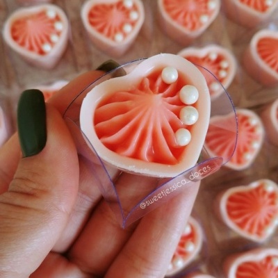 Bombom em forma de coração com decoração rosa e pérolas brancas segurado por mão com unhas verdes.