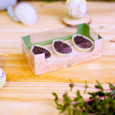 Três ovos de chocolate branco com cobertura de chocolate preto em caixa transparente sobre mesa de madeira