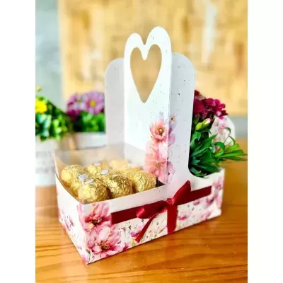 Cesta decorativa com chocolates dourados e flores artificiais, padrão floral e laço vermelho.