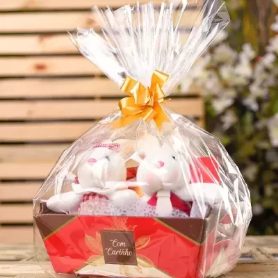 Cesta de presente com dois coelhos de pelúcia e laço laranja, em fundo de ripas de madeira e flores.