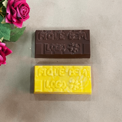 Dois tabletes de chocolate castanho escuro e amarelo com texto e símbolo de flor em relevo sobre superfície cinzenta e flores rosas ao lado