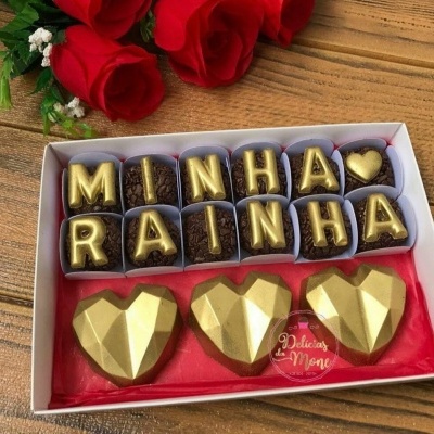Bombons com letras douradas em caixa com 3 bombons dourados em forma de coração e rosas vermelhas ao fundo
