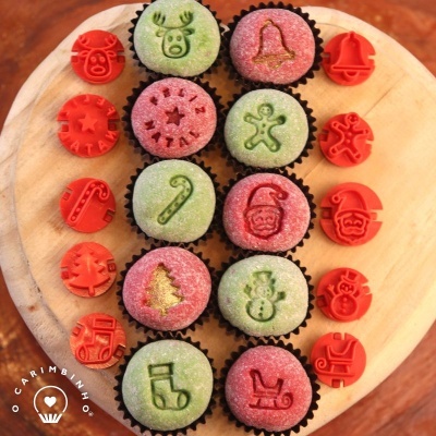 Doze cupcakes decorados com desenhos natalícios em glacê verde e rosa, num prato de madeira, rodeados por carimbos vermelhos com motivos natalícios.