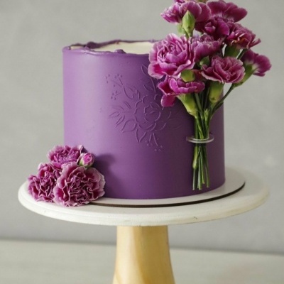 Bolo roxo decorado com flores naturais cor-de-rosa num suporte branco com base em madeira.