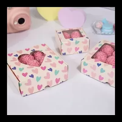 Caixas de papel com coração recortado, doces rosados dentro, superfície branca e fundo desfocado com itens coloridos.