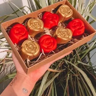 Caixa com sabonetes em forma de rosas vermelhas e douradas segura por uma mão em frente a planta verde