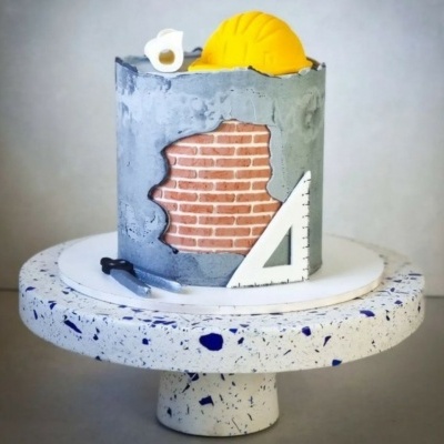 Bolo decorado com tema de construção com capacete amarelo e ferramentas em miniatura, sobre suporte branco com manchas azuis