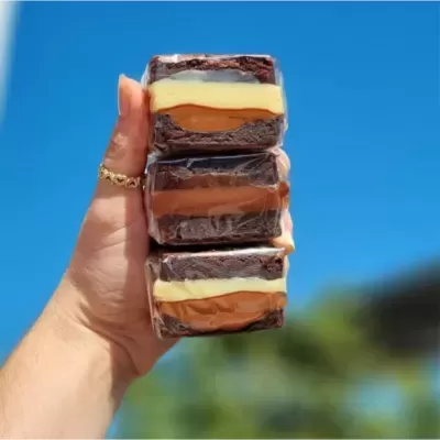 Três bolos tipo brownie com camadas de chocolate e creme, segurados por mão diante de céu azul.