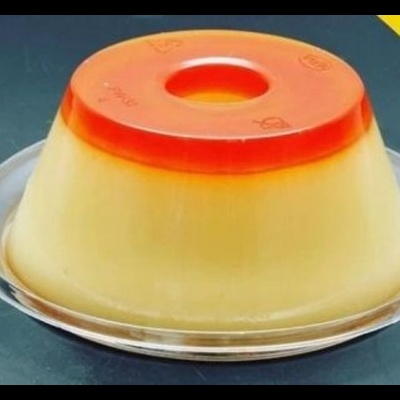 Flan duas camadas amarelo e vermelho num prato transparente sobre fundo escuro com triângulo amarelo.