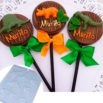Pirulitos decorativos com dinossauros e nome Murilo, com laços coloridos, sobre papel rendilhado, e molde de silicone azul com formas de dinossauros.