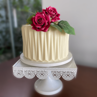 Bolo branco com padrão geométrico e decoração de rosas vermelhas num suporte branco rendilhado.