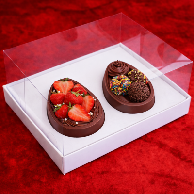 Duas metades de ovo de chocolate decoradas com morangos e brigadeiros, em caixa branca com tampa transparente