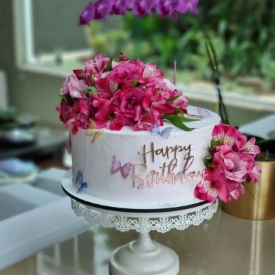 Bolo branco decorado com flores cor-de-rosa e texto Happy Birthday dourado