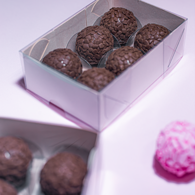 Caixa transparente com seis bombons de chocolate escuro e um bombom rosa desfocado ao lado