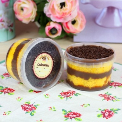 Dois bolos em taças transparentes com camadas de creme amarelo e chocolate, cobertura de chocolate granulado, etiqueta 'Cakepedia', sobre pano floral branco