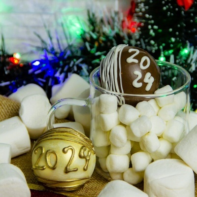 Bolas de Natal decorativas com número 2024 entre marshmallows brancos numa caneca de vidro, com decoração natalícia colorida ao fundo.