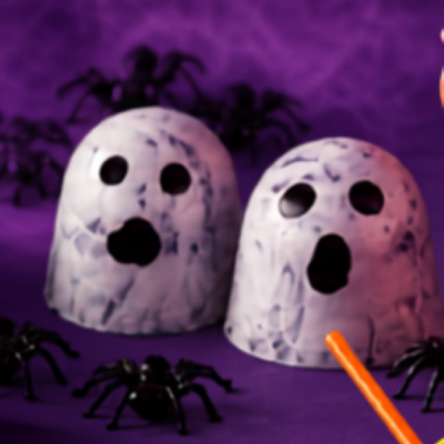 Dois copos brancos marmoreados com olhos e boca pretos, simulando fantasmas, com aranhas pretas ao redor sobre fundo roxo.