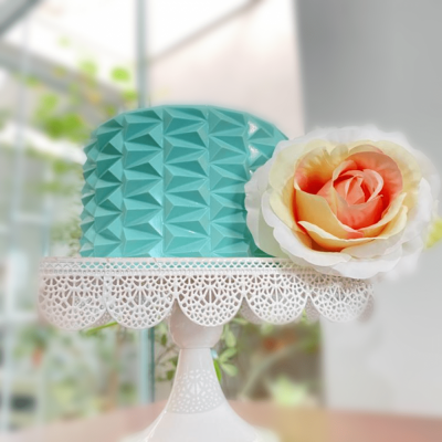 Bolo azul turquesa com textura geométrica e flor artificial num suporte branco rendado