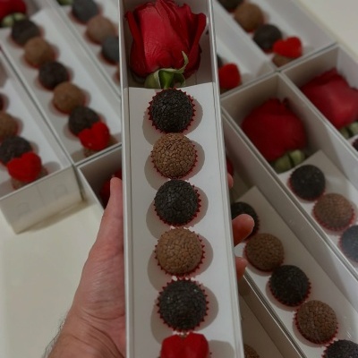 Caixa comprida branca com rosa vermelha e bombons variados em confeitos preto, castanho e vermelho em forma de coração.