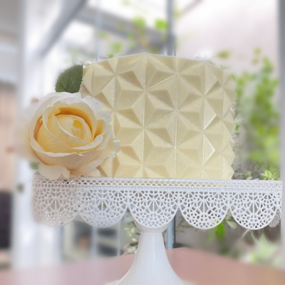 Bolo quadrado amarelo claro com padrão geométrico e flor decorativa numa base branca rendada