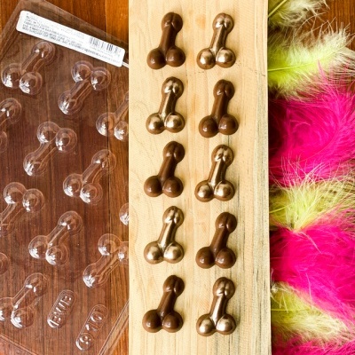 Bombons em forma de pênis em chocolate castanho e dourado sobre tabuleiro de madeira com decoração preta, embalagem plástica e penas coloridas ao lado.