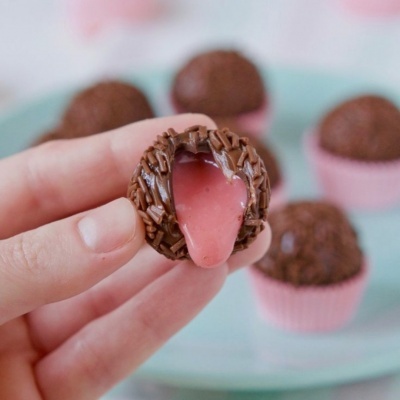 Doce brigadeiro de chocolate com recheio rosa cremoso segurado por mão e outros brigadeiros no prato verde-claro ao fundo