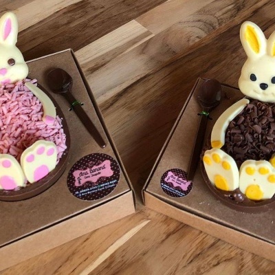 Dois ovos de chocolate com coelhos de chocolate branco e colheres, em caixas de papelão castanho.