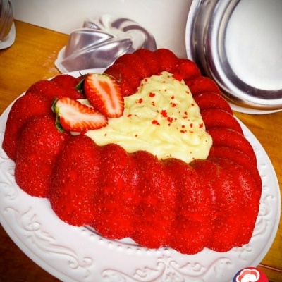 Bolo vermelho com cobertura branca e morangos num prato branco com formas metálicas ao fundo