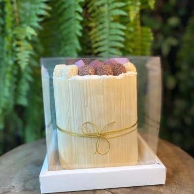 Bolo cilíndrico com brigadeiros variados no topo em embalagem transparente sobre base branca