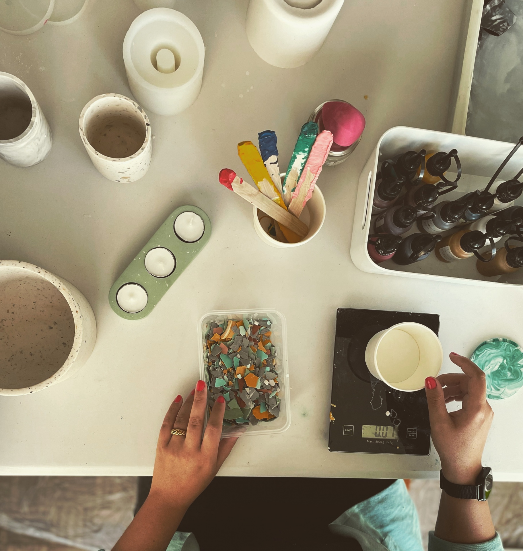 Mesa de trabalho com potes de cerâmica, palitos coloridos, balança digital, caixa com pedaços de cerâmica e mãos femininas