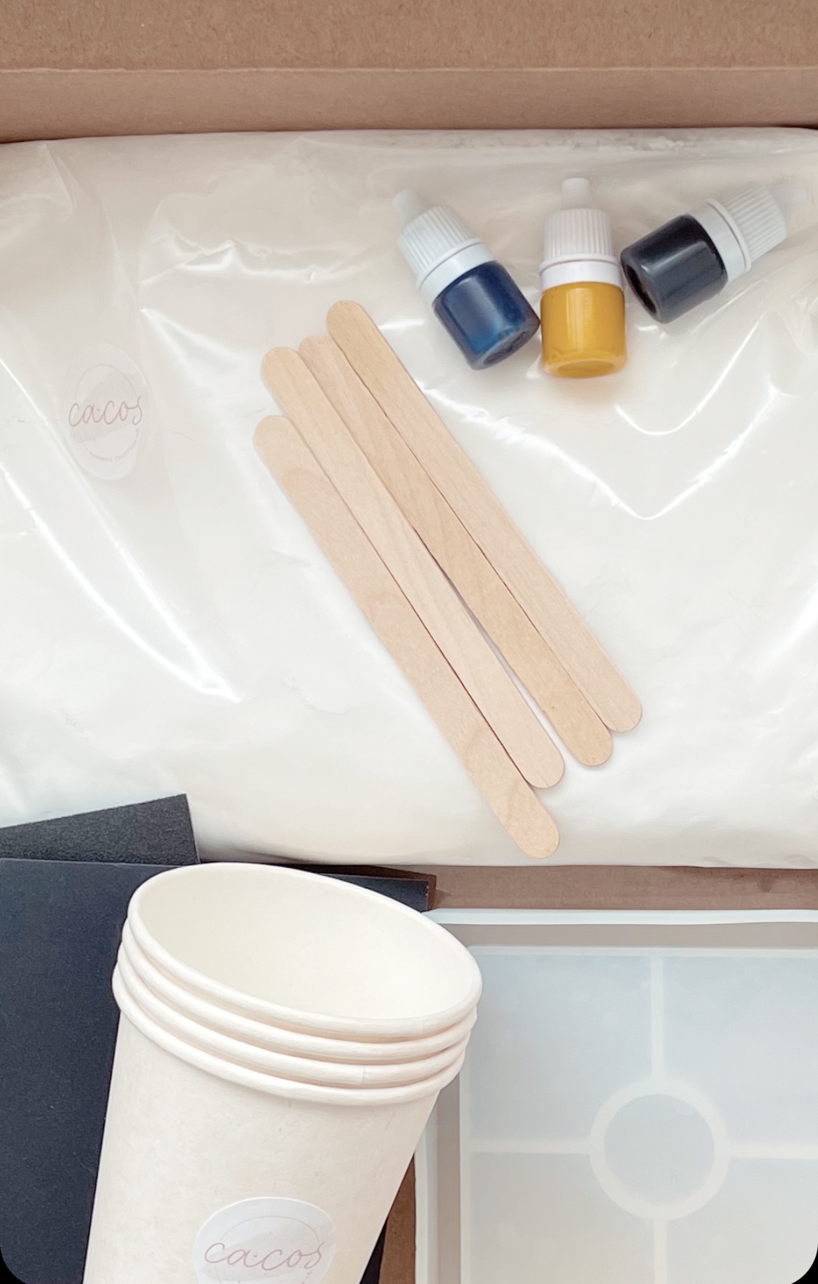 Material de artesanato com palitos de madeira, tinta azul, amarelo e preto, copos de papel e folhas de papel, em caixa de cartão com selo 'cacos'.