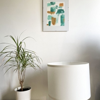 Quadro abstrato verde e bege, planta em vaso branco, candeeiro de mesa com base de madeira e cúpula branca