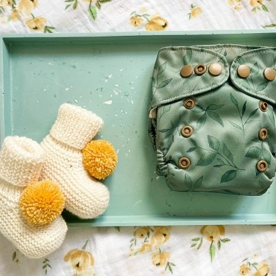 Fralda reutilizável verde com padrão de folhas e luvas de malha creme com pompons amarelos numa bandeja verde