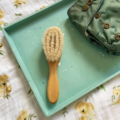 Escova de cabelo com cabo de madeira e fralda reutilizável verde numa bandeja azul clara sobre tecido floral branco