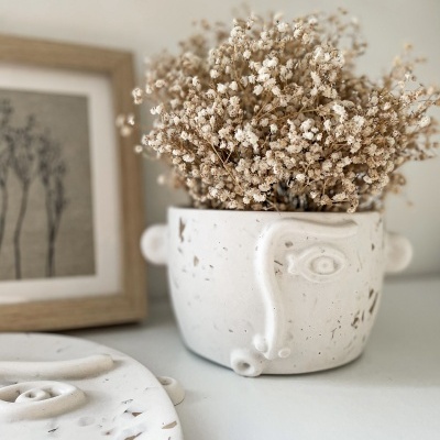 Vaso de cerâmica branco com rosto abstrato e flores secas, em superfície branca próximo de quadro com plantas.