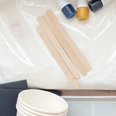 Material de artesanato com palitos de madeira, tinta azul, amarelo e preto, copos de papel e folhas de papel, em caixa de cartão com selo 'cacos'.