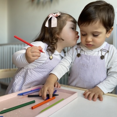 Duas crianças pequenas com macacões lilás às riscas e camisolas brancas, com lápis de cor numa bandeja rosa numa mesa branca num quarto com decoração suave.