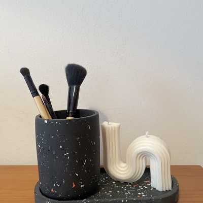 Porta-pincéis preto com manchas coloridas contendo pincéis de maquiagem e vela branca ondulada sobre base preta