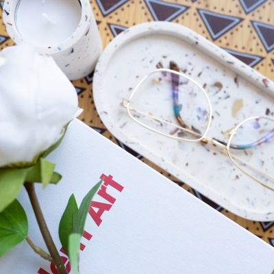 Mesa com vela, vaso com rosa branca, manta branca, suporte oval branco com óculos dourados e padrão geométrico