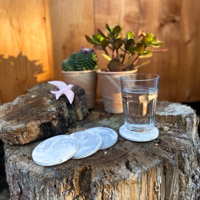 Bases para copos brancas marmoreadas sobre tronco de madeira com copo de água e plantas