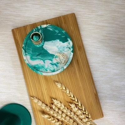 Base redonda de resina verde-azulada e branca com acessórios sobre tábua de madeira e espigas de trigo num fundo bege