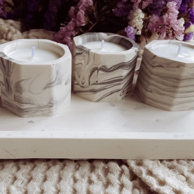 Três velas em recipientes marmorizados sobre bandeja com flores ao fundo