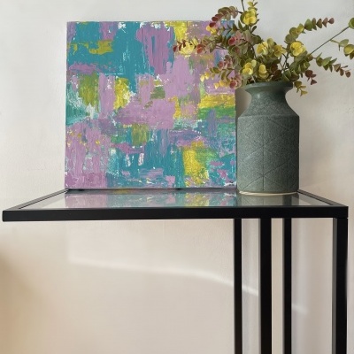 Mesa de canto em vidro e metal com vaso cinza e flores artificiais ao lado de pintura abstrata colorida