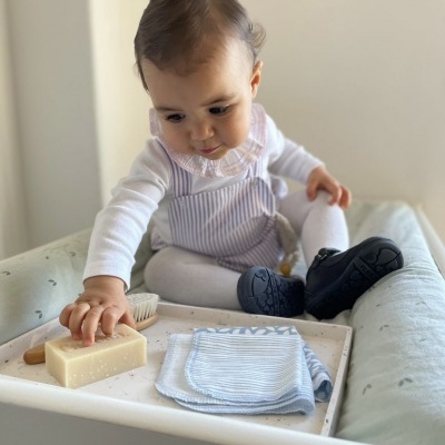 Bebé com roupa às riscas sentado num berço com sabonete, escova e pano