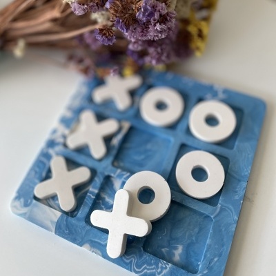 Jogo da velha com peças brancas em tabuleiro azul marmoreado e flores secas ao fundo.