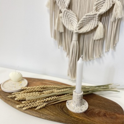 Suporte para vela com vela branca, trigo seco, sabonete em formato de concha sobre base de madeira com decoração macramê na parede.
